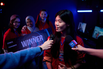 Asian teenage girl gamer being interviewed by reporters after esports tournament victory, standing in front of diverse teenage girls team holding winner sign, gaming monitors in background © pressmaster