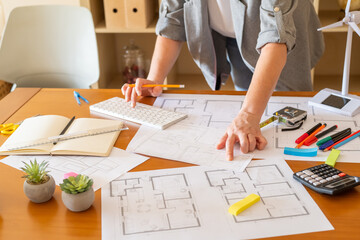 Architect working on a sustainable building project is examining blueprints and pointing at a specific detail on the plan, with a miniature wind turbine and solar panel placed on the desk