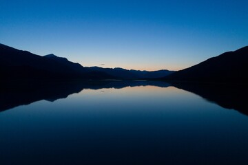 Silhouetted peaks are casting reflection on lake water and dusky sky is fading in flat design