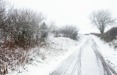 Flat design is showing winding snow-covered lane curving uphill with twin tracks and hedgerows