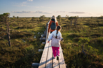 Mother and daughter walk along a wooden path through a picturesque bog at sunset, enjoying a hike...