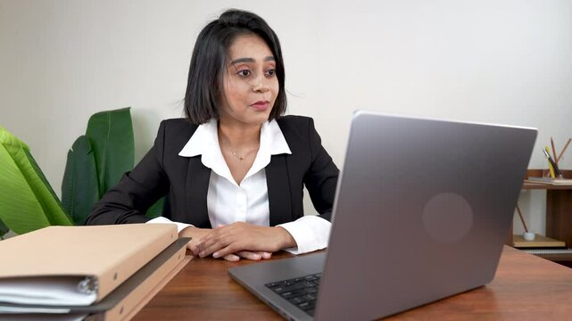 Close-up of surprised astonished attractive young Indian businesswoman sits on chair at workplace desk look at laptop screen using internet chatting on videocall nod head to agree with amazed face.