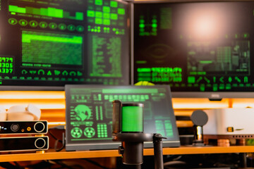 A close-up of a black microphone with green LED lighting, placed in front of multiple computer screens displaying hacker-style green codes and data in a dark modern workspace.