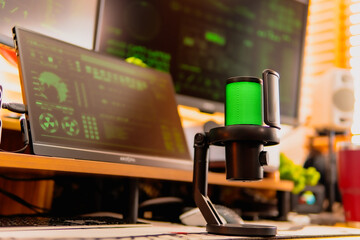 A close-up of a black microphone with green LED lighting, placed in front of multiple computer screens displaying hacker-style green codes and data in a dark modern workspace.