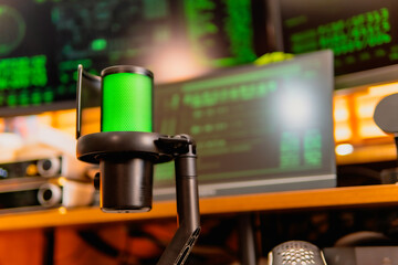 A close-up of a black microphone with green LED lighting, placed in front of multiple computer screens displaying hacker-style green codes and data in a dark modern workspace.