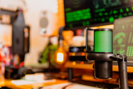 A close-up of a black microphone with green LED lighting, placed in front of multiple computer screens displaying hacker-style green codes and data in a dark modern workspace.