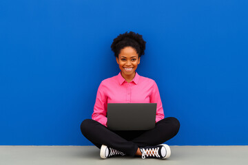 A smiling woman sits on the floor with a laptop against a blue wall