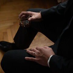 A man in a suit sits and holds a glass of amber liquid, lost in thought.