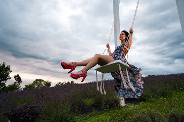 Woman in a floral dress enjoys a swing ride overlooking a lavender field under a cloudy sky.
