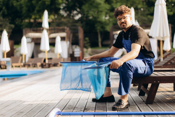 Pool cleaner holding cleaning nets sitting on sunbed near swimming pool