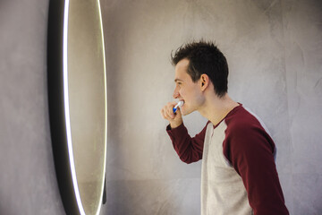 Young man practicing daily oral hygiene, brushing his teeth in front of a round illuminated mirror...