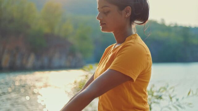 Young Indian woman sitting in padmasana pose on yoga mat near a lake, morning sunlight reflecting on water surface.