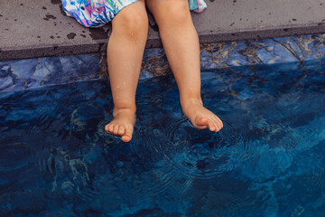 young girl sits by swimming pool, dipping her feet into the water.