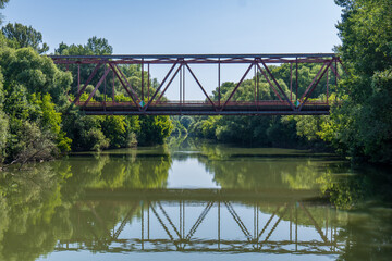 Metal bridge over the Bodrog river in Viničky village, Slovakia. Green trees on both sides and calm water reflecting the bridge structure on a sunny summer day.
