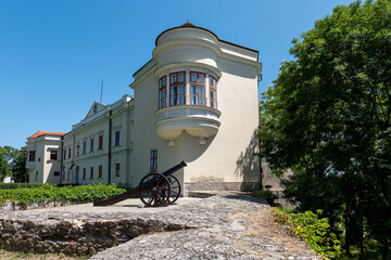 Historic Sárospatak Castle in Hungary with old black cannon on stone terrace, Renaissance architecture, green trees, and clear blue sky.