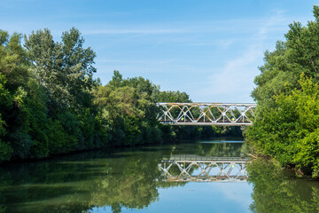 Railway bridge over Bodrog river surrounded by green trees. Bridge and trees reflected in calm water on a sunny summer day.