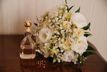 A wedding bouquet of white roses lies on the table next to two gold wedding rings for the bride and groom and a bottle of perfume stands next to them on their wedding day
