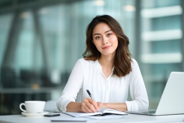 Obraz premium A young woman in a white blouse sits at a desk, writing in a notebook with a laptop and coffee cup nearby in a bright, modern office setting.