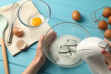 Woman whisking egg whites with hand mixer at light blue wooden table, above view