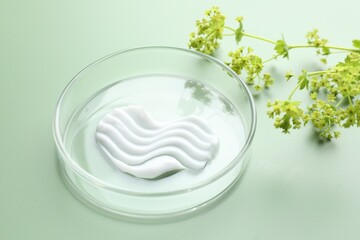 Cosmetic product in Petri dish and floral decor on green background, closeup