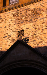 An ancient stone cross adorning the top of the Abbey of Sainte-Foy casts its shadow on the medieval stone facade illuminated by the golden light of sunset, in Conques, France.