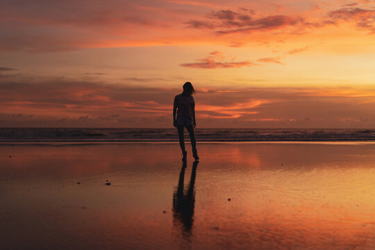 Young woman with arms raised facing colorful sunset at low tide beach - Powered by Adobe