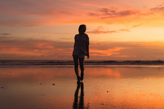 Young woman with arms raised facing colorful sunset at low tide beach - Powered by Adobe