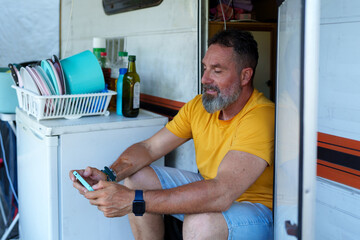 Mature traveler relaxing inside camper doorway, playing smartphone game during sunny afternoon outdoors
