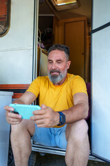 Senior male playing mobile game, relaxing near camper van entrance
