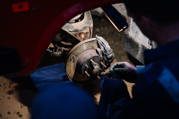 A mechanic works on a rusty car brake disc and suspension system under a wheel arch, using gloves...
