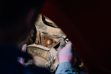 Mechanic working on a rusty car suspension system, focusing on bolts and brake components under the wheel arch in close-up view.