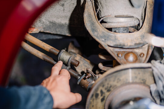 Mechanic’s hand pointing at a worn car suspension bushing and rusted linkage near the wheel assembly during a vehicle inspection or repair process. - Powered by Adobe