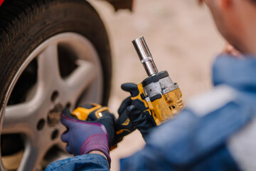 Close-up of a mechanic using a yellow power tool to tighten or loosen lug nuts on a car wheel while wearing protective gloves and a work uniform.