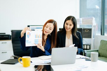 Two Asian businesswomen in suits analyze reports together at a modern office desk. A scene of...