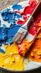 Close-up of textured abstract painting with bold strokes in blue, teal, red, and orange. A paintbrush lies diagonally, stained with color, highlighting vibrant process and texture.

