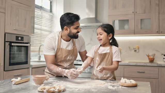 A father and daughter bake together in a modern kitchen. The father is a young South Asian man with a beard. The daughter is a young South Asian girl with long hair. Flour is scattered on the counter.