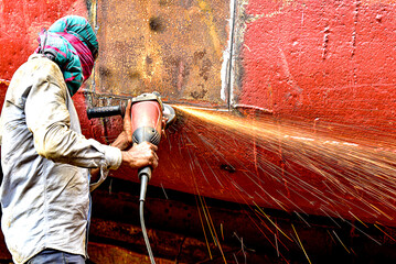 Worker grinding metal in shipyard during daytime while sparks fly in coastal setting