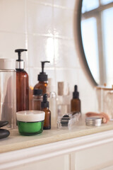 Assortment of skincare products including glass bottles, pump dispensers, cream jar, facial roller arranged on bathroom shelf reflecting daily beauty and skincare routine essentials