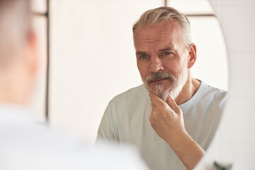 Middle aged Caucasian man touching beard while looking at own reflection in mirror, practicing skincare routine, showing thoughtful facial expression in bathroom setting