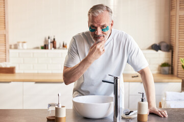 Middle aged Caucasian man standing at bathroom sink applying skincare routine with under eye patches, touching chin thoughtfully, engaging in personal grooming and self care