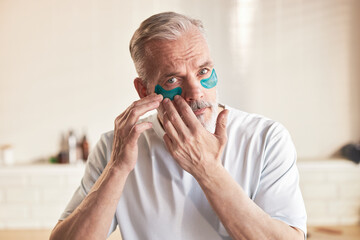 Portrait of middle aged Caucasian man applying under eye patches to face while looking into camera, demonstrating skincare routine and self care in bathroom setting