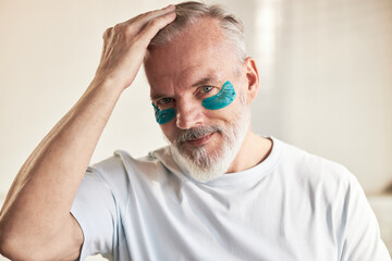 Portrait of middle aged Caucasian man smiling at camera, applying blue under eye patches, touching...