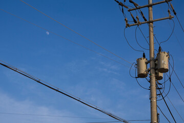 Electric poles on the street, transformers and wires hanging from them, and half a moon floating in...
