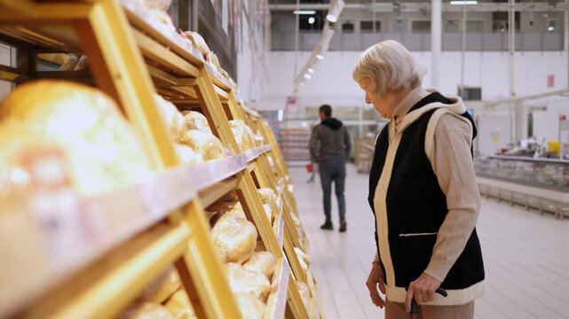 An old grandmother with gray hair chooses and buys fresh bread in a large supermarket. The pensioner chooses bread.