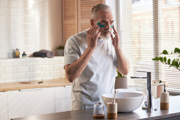 Middle aged Caucasian man applying under eye patches while standing at bathroom sink, focusing on...