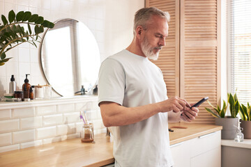 Middle aged Caucasian man standing in modern bathroom holding smartphone, preparing for skincare routine with grooming products and plants visible on countertop