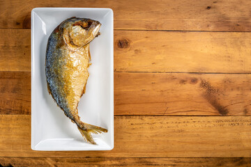 Top view of crispy fried mackerel on a white  plate placed on a natural wooden surface. Thai cuisine presentation perfect for menu or cooking content.