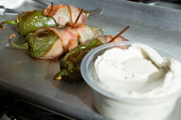 A view of some bacon wrapped jalapeno poppers next to a container of herbal cream cheese.