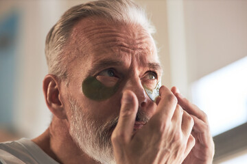 Middle aged Caucasian man applying under eye patches during skincare routine, gently pressing...