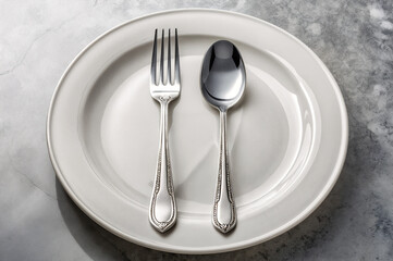 shows a white, oval-shaped plate with a fork and a spoon placed on it, both made of shiny metal with intricate designs along the handles. The plate is on a textured, grey surface.